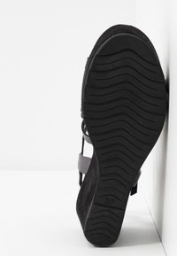 Semelle noire de sandale avec motif ondulé, taille 37, inclinée sur fond blanc montrant une partie des brides supérieures.