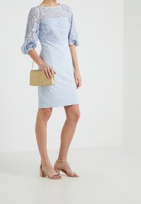Jasnoniebieska sukienka z koronkową górą, krótkimi bufkami i dopasowaną spódnicą. Złota torebka crossbody o pikowanej fakturze i cieliste sandały na obcasie.