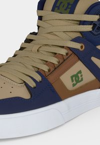 Basket montante avec lacets beiges, panneaux beige et marine, logo DC vert et semelle blanche.