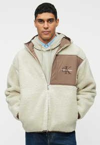 Casaco de fleece em creme com um painel castanho e logotipo. Apresenta um fecho éclair, capuz e punhos canelados. Estilizado com um sweatshirt leve por baixo.
