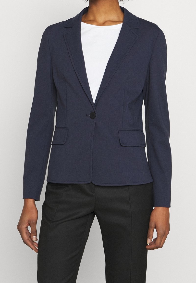 Femme portant un blazer bleu marine à un bouton sur une chemise blanche avec un pantalon noir, vue du cou jusqu'aux hanches sur un fond uni.