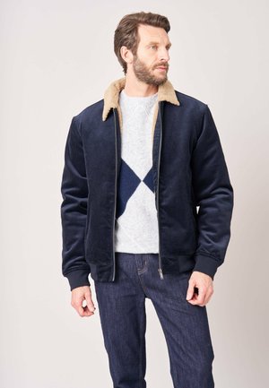 Man draagt een marineblauwe ribfluwelen jas met een beige shearlingkraag, een grijs en marineblauw gebreide trui met patroon en donkerblauwe spijkerbroek, kijkt zijwaarts.