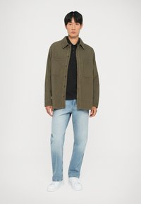 Giacca in corduroy verde oliva con due tasche sul petto, indossata sopra una polo nera, abbinata a jeans azzurri chiari e sneakers bianche.