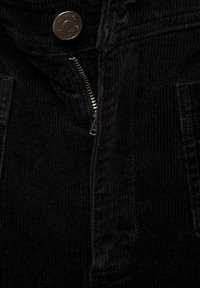 Tessuto in corduroy nero con texture a coste, dotato di un bottone a pressione in metallo e di una zip. Include dettagli con tasche sul davanti.