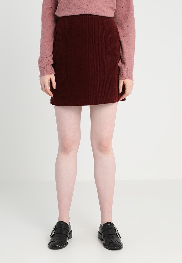 Personne portant une jupe en velours côtelé bordeaux, un pull rose et des mocassins noirs à boucle, debout devant un fond blanc uni.