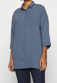 Chemise bleue surdimensionnée en tissu froissé avec une fermeture à boutons, un col, des manches trois-quarts et une poche poitrine.