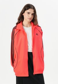 Ljusrosa sportjacka med svarta ränder, dragkedja och ståkrage. Lättviktig, mjuk textur med Adidas-logotyp på bröstet.