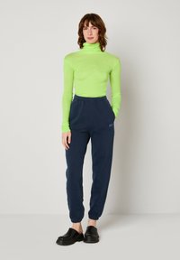 Un turtleneck vert fluo à manches longues, un pantalon de jogging marine avec taille élastique et des chaussures noires à enfiler ; un design simple et une silhouette ajustée.