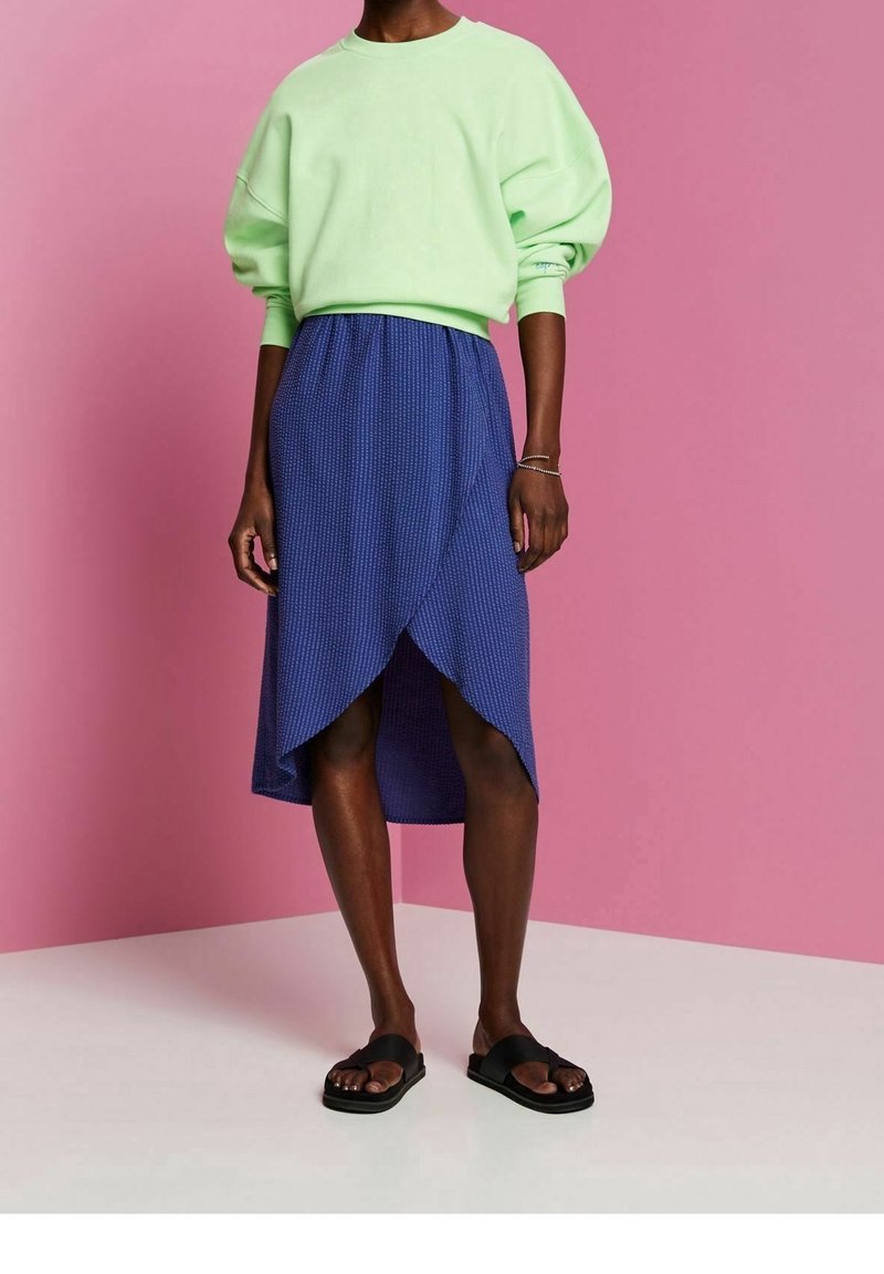 Personne portant un sweat-shirt ample vert clair, une jupe portefeuille bleue texturée, des sandales noires à bouts ouverts, debout contre un mur rose et un sol blanc.