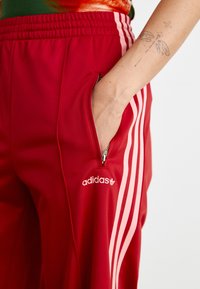 Sarkani sporta bikses ar rozā sānu strīpām, aprīkotas ar rāvējslēdzēja kabatu un baltu "adidas" logotipu uz kreisā augšstilba. Gluda tekstūra.