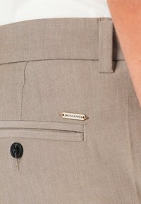 Beige bukser har et slankt design med en subtil stribet tekstur, en sort knap og et guldmærke præget med "JACK & JONES."