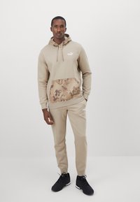 Conjunto de sudadera beige y joggers con un bolsillo frontal de color marrón con patrón, que presenta un logo blanco y capucha con cordón, combinado con zapatillas deportivas negras.