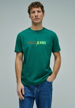 Homme aux cheveux bouclés portant un t-shirt vert avec le texte "Salsa/Jeans" et un jean bleu, debout devant un fond gris uni.