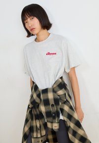 Šedé bavlněné tričko s červeným logem "ellesse". Černá a béžová kostkovaná flanelová košile uvázaná v pase. Jednoduchý, neformální design.