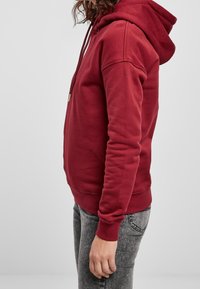 Sudadera roja con capucha y cordón, puños y dobladillo acanalados. Vista lateral que muestra un ajuste holgado. Combinada con jeans grises, sobre un fondo neutro.