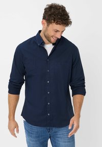 Marineblauw overhemd met knopen, gemaakt van katoen, met een borstzak, knoopmanchetten en een gebogen zoom. G gedragen met een wit T-shirt eronder.