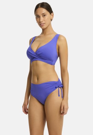 A woman poses confidently in a vibrant purple bikini, featuring a wrap-style top and drawstring bottoms, against a plain white background.