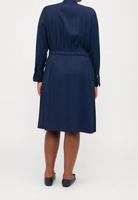 Marineblaues, knielanges Kleid mit tailliertem Schnitt, langen Ärmeln und geknöpften Manschetten, aus glattem Stoff mit minimalistischer Gestaltung.