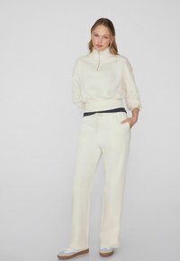 Crème zip-nek sweat-top met brede mouwen, gecombineerd met bijpassende crème joggers met een marineblauwe trekkoord en gebandeerde taille. Witte sneakers maken de look af.
