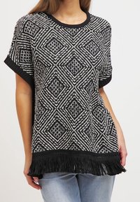 Haut en tricot à motifs noir et blanc avec un design en losanges, manches courtes, col côtelé et ourlet frangé. Porté sur un denim clair.