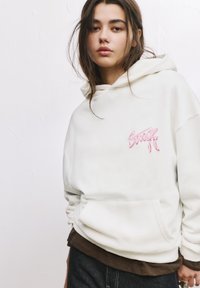 Hvid hættebluse med pink broderet design på brystet, udstyret med en kengurulomme og ribbede manchetter. Laget over en brun top.