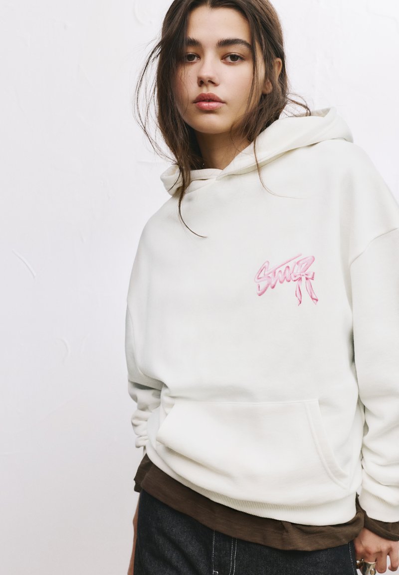 Hvid hættebluse med pink broderet design på brystet, udstyret med en kengurulomme og ribbede manchetter. Laget over en brun top.