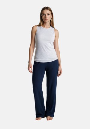 Kobieta stojąca boso, ubrana w bezrękawnik w kolorze białym oraz luźne, ciemnoniebieskie spodnie na jednolitym jasnym tle.