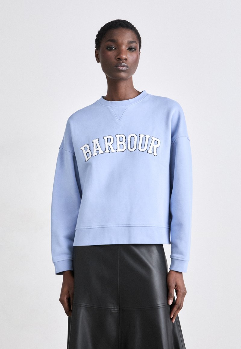 Világoskék pulóver, amelyen fehér hímzéssel szerepel a "BARBOUR" felirat, kerek nyakkivágással, esősen esőn vállrésszel és laza szabással. Fekete bőr szoknyával párosítva.