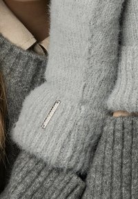 Szürke kötött pulóver bordás textúrával; puha, bolyhos nyakkal és egy ezüst fém címkével a mandzsettán.