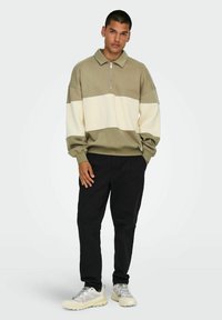 Khaki and cream color-blocked zip-up pullover with a collar, paired with black pants and light grey shoes. Textured fabric, relaxed fit.