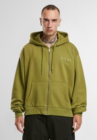 Olive krāsas zip-up kapuce, izgatavota no mīksta auduma, ar kangaroo kabatu un ribotām piedurknēm, kurā uz krūtīm ir tonāls logotips.