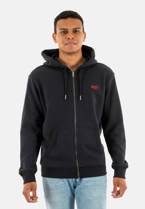 Marine blauwe zip-up hoodie van zacht materiaal, met een voorzak, koordjes en een klein rood logo op de borst. Gekocht met blauwe jeans.