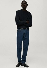 Pulover negru, blugi albaștri și pantofi negri. Blugii sunt largi, cu o textură tipică de denim. Design simplu, fără modele vizibile.