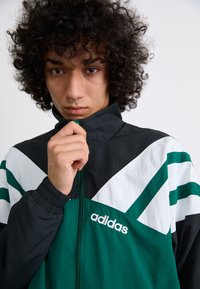 Black, green, and white jacket with a zip front, featuring bold color blocking, stripes, and an "Adidas" logo on the chest. Textured fabric.