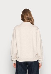 Sweat-shirt beige avec col à revers, poignets et ourlet côtelés. Le tissu semble doux et texturé, avec une coupe décontractée et un design minimaliste.