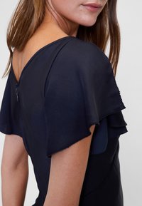 Tengerészkék ruha rövid, rétegezett hullámos ujjakkal és V-alakú hátrésszel. Sima textúra és finom varrással díszített részlet. Cipzáras záródás hátul.