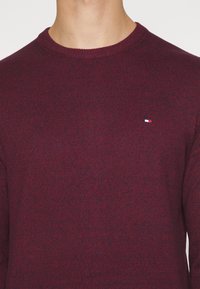 Bordeauxroter gestrickter Pullover mit Rundhalsausschnitt und gerippten Kanten. Kleines Logo auf der Brust in Blau, Weiß und Rot. Weiche Textur und figurbetontes Design.