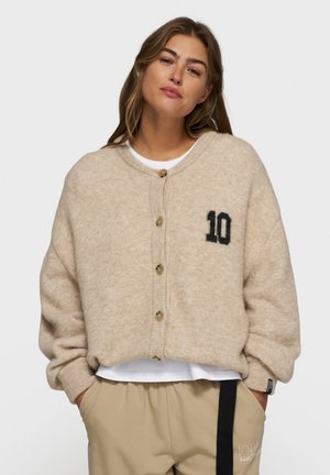 Junge Frau in einem beigen, knopfbelegten Cardigan mit der Zahl "10" auf der Brust und beigen Hosen mit einem schwarzen Streifen, die Hände in den Taschen, neutraler Hintergrund.