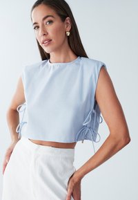 Lyseblå cropped top med rund halsudskæring og korte ærmer, med sidelukninger med guld accenter. Glat tekstur, afslappet design.