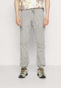 Persoană îmbrăcată cu pantaloni cargo gri deschis, cu talie elastică și manșete elastice, asortați cu încălțări de drumeție verzi și maronii.