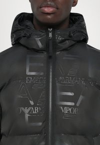Schwarze Daunenjacke mit glänzender Oberfläche, ausgestattet mit einer Kapuze, Reißverschluss und geprägten Logos. Texturiert mit modernem Design.