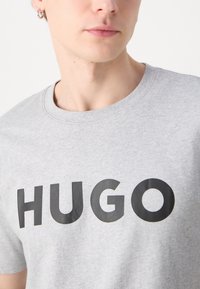 Pelēks kokvilnas T-krekls ar apaļu kaklu un īsām piedurknēm. Ar lielu melnu "HUGO" tekstu uz krūtīm. Gluda tekstūra, ikdienas piegriezums.