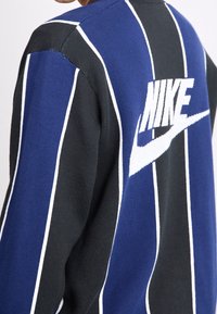 Musta ja sininen raidallinen neulepaita, jossa on selkeä valkoinen Nike-logo takana. Valmistettu teksturoitusta kankaasta, jossa on kontrastivärejä.