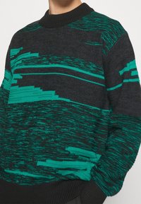 Pull tricoté gris foncé avec des motifs abstraits turquoise vif. Il présente un col rond côtelé et des manches longues avec une finition texturée.