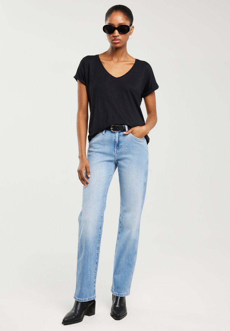 Femeie purtând tricou negru cu decolteu în V, blugi bleu deschis cu curea neagră, cizme negre cu toc și ochelari de soare negri, stând pe un fundal simplu.