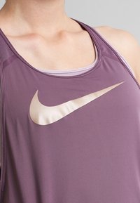 Canottiera sportiva viola con una texture leggera e liscia. Presenta un grande logo Nike dorato, scollatura tonda e design a corsetto.