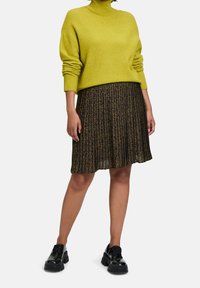 Frau trägt einen limettengrünen Rollkragenpullover, einen plissierten Leopardenmuster-Rock und schwarze klobige Plateau-Schuhe, steht vor weißem Hintergrund.