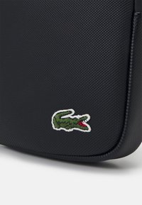 Black textured bag featuring a green embroidered crocodile logo. The material has a geometric pattern and a smooth, rounded edge.