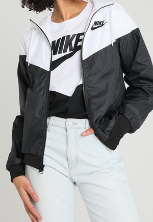 Osoba ubrana w czarno-białą kurtkę typu wiatrówka Nike na białym t-shircie Nike i jasnoniebieskich dżinsach, ręka w kieszeni.