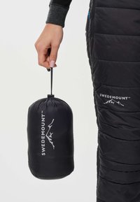 Ręka trzyma czarny woreczek Swedemount ze ściągaczem, obok czarnych pikowanych spodni z logo Swedemount na udzie.